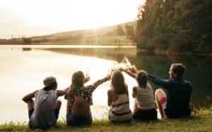 ragazzi in viaggio che brindano con bottiglie davanti a un lago al tramonto